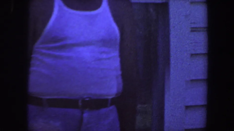 Adult man in a white tank top stands near house siding in a blue-purple 1959 Iowa 8mm home-movie frame.