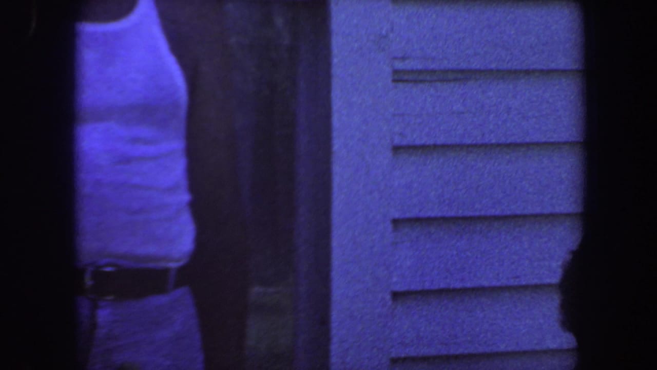 Adult in a white sleeveless shirt standing beside a house with horizontal siding.