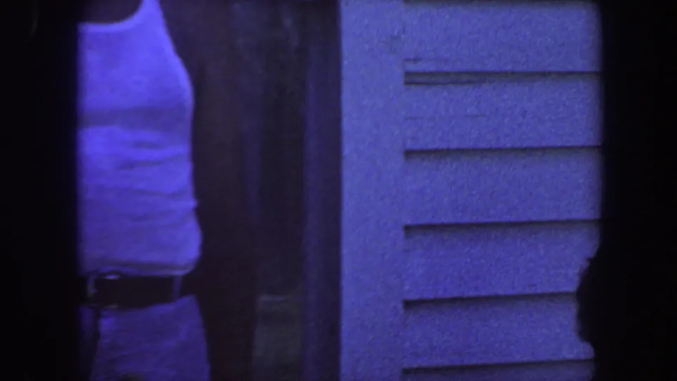 In 1959 Iowa home-movie footage, an adult in a white sleeveless shirt stands near horizontal house siding with a strong blue film cast.