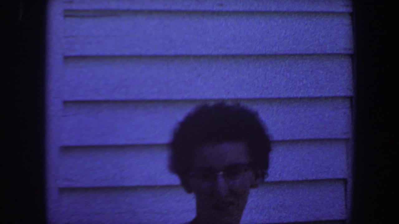 An adult with short dark hair and glasses faces the camera outside a house in Iowa, 1959, with blue-tinted 8mm film grain.