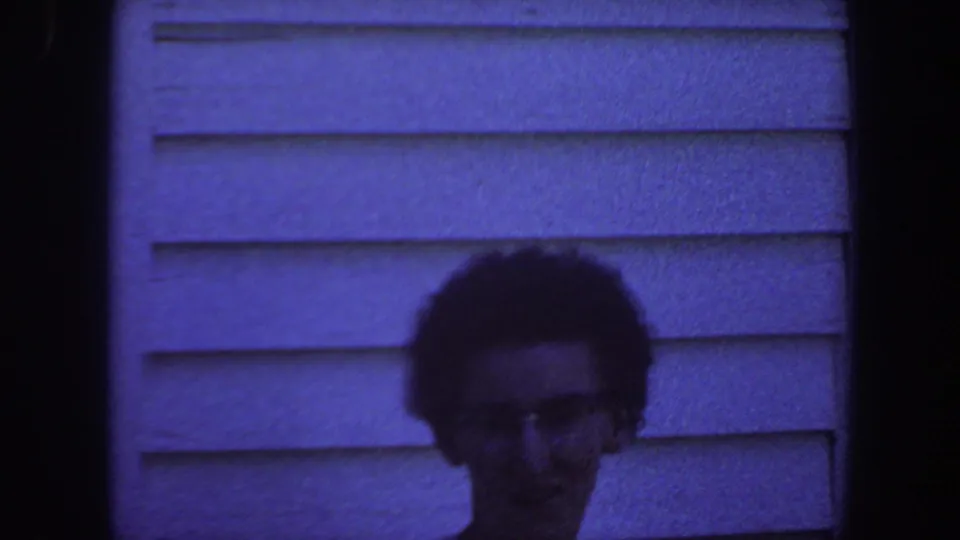 An adult with short dark hair and glasses faces the camera outside a house in Iowa, 1959, with blue-tinted 8mm film grain.