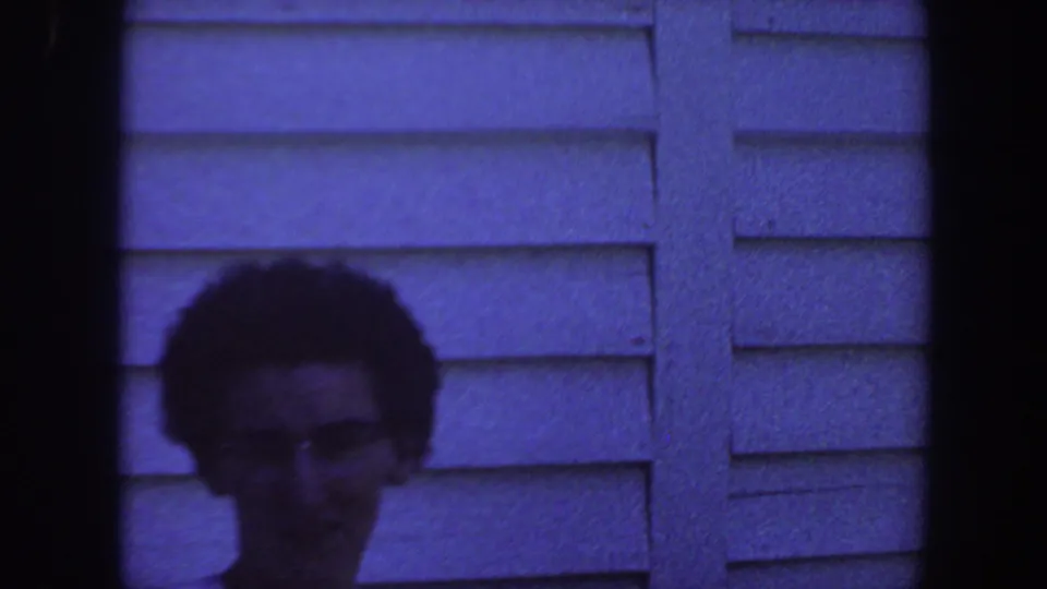 A partially framed adult with glasses appears low in the shot against a 1959 Iowa house exterior with horizontal siding.