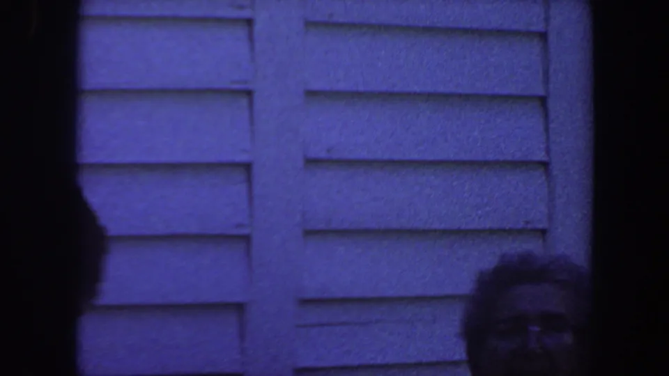 A partly visible adult with glasses appears at the lower right in blue-tinted 8mm footage of an Iowa house exterior from 1959.