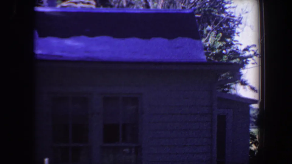 A closer 8mm view of the white house exterior shows windows, siding, shrubs, and shaded yard under a strong purple cast.