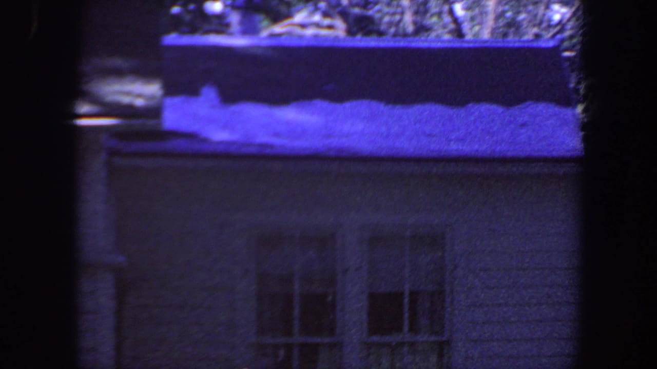 1959 Iowa 8mm frame showing a modest house wall, window, gutter, and snow patches on the shingled roof under a blue-purple cast.