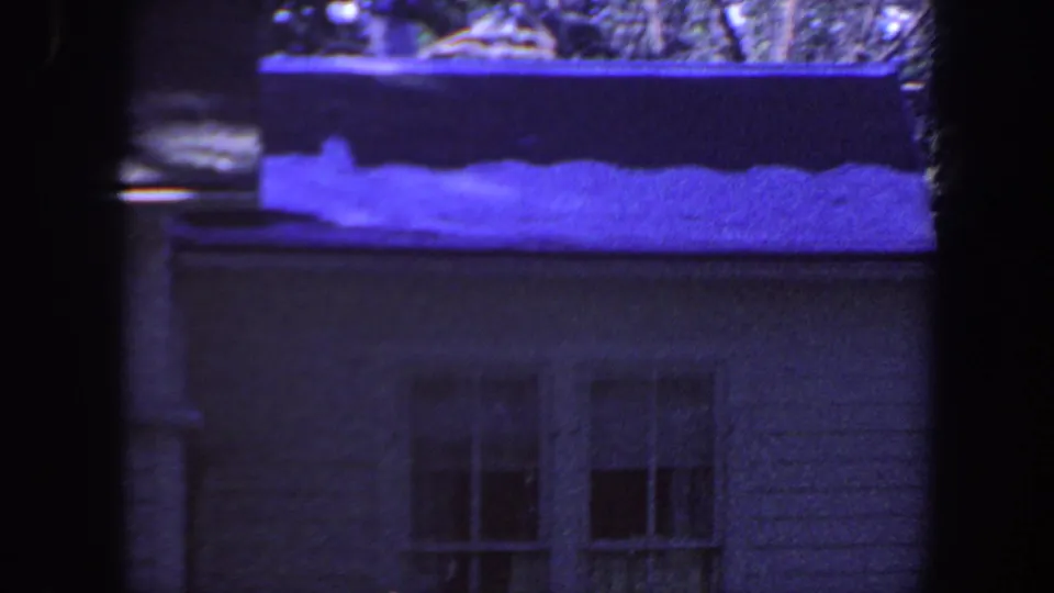 1959 Iowa 8mm frame showing a modest house wall, window, gutter, and snow patches on the shingled roof under a blue-purple cast.