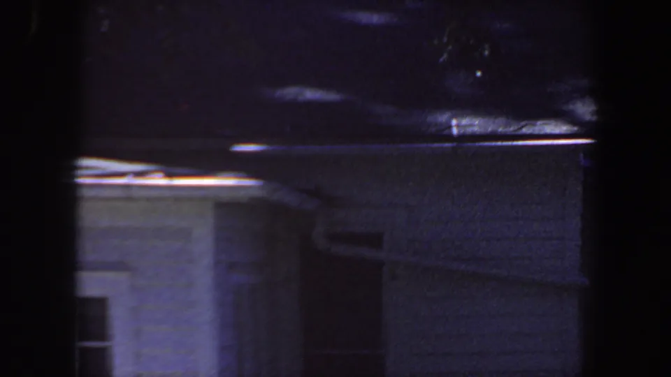 1959 Iowa frame with a shadowed house wall, roof edge, gutter line, and partial window, tinted blue-purple by aged 8mm film.