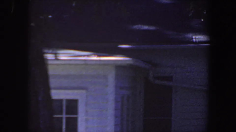 1959 Iowa 8mm frame of a white house exterior at night, with a divided window and dark tree trunks crossing the view.