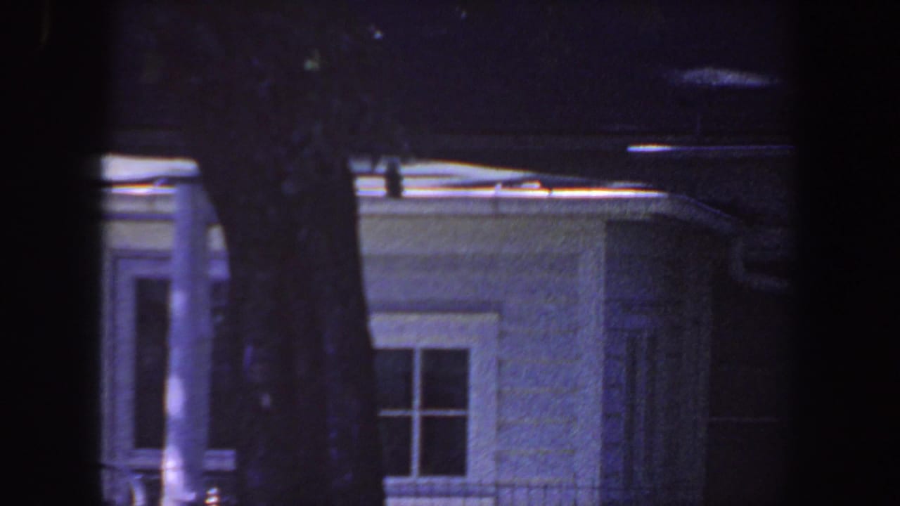 1959 Iowa home-movie frame showing a white residential exterior with roofline, doorway detail, window, and a large tree trunk.