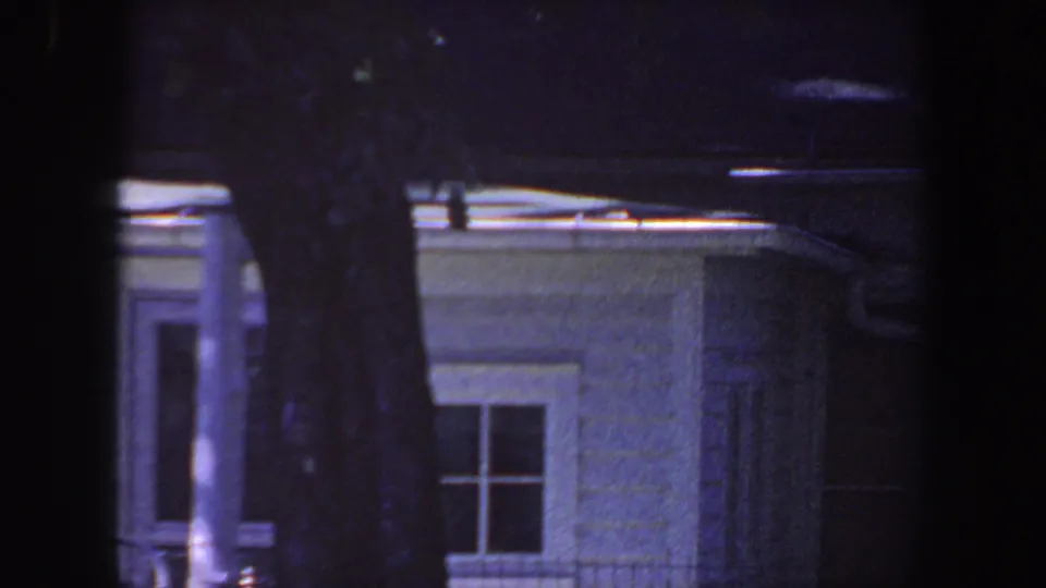 1959 Iowa home-movie frame showing a white residential exterior with roofline, doorway detail, window, and a large tree trunk.