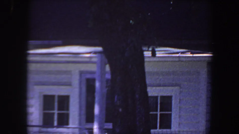 1959 Iowa 8mm frame with white siding, two window areas, roof gutter, and a large foreground tree trunk in low light.