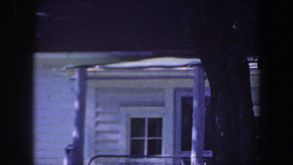 1959 Iowa home-movie frame showing a white clapboard house, a divided window, doorway detail, and a dark tree trunk at night.