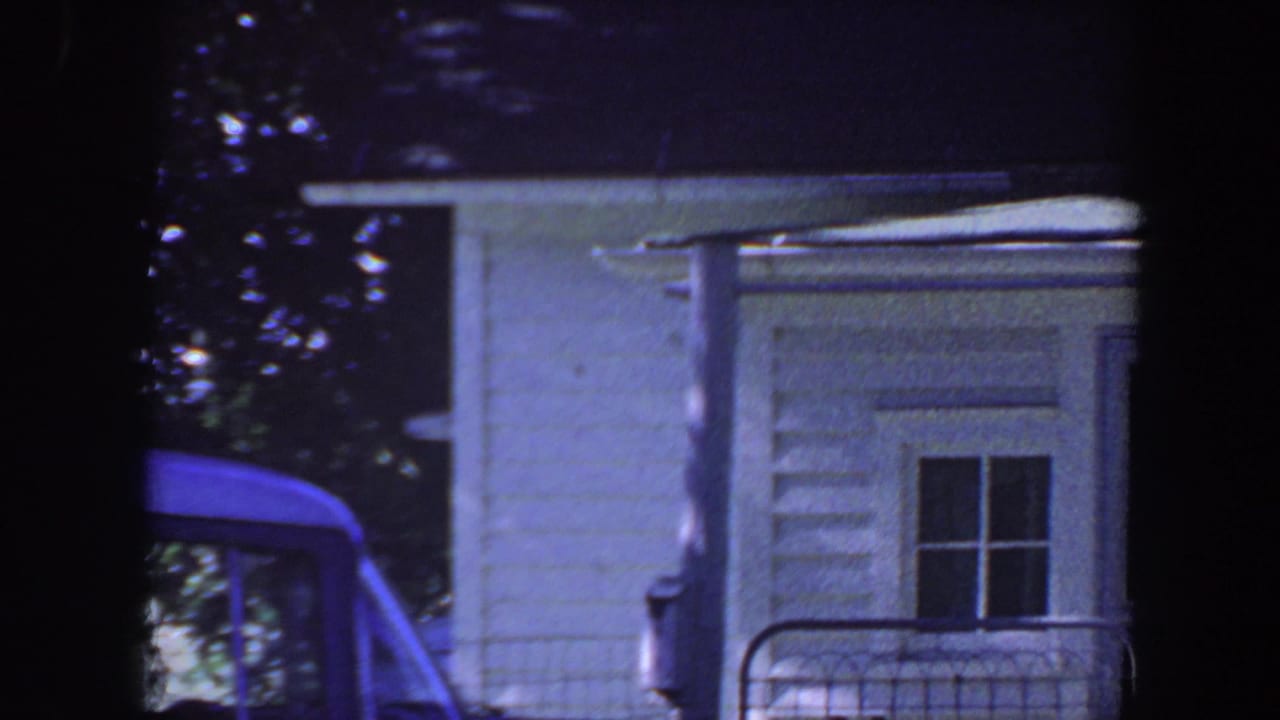 1959 Iowa home-movie frame showing a white house, porch railing, leafy trees, and part of a blue car.