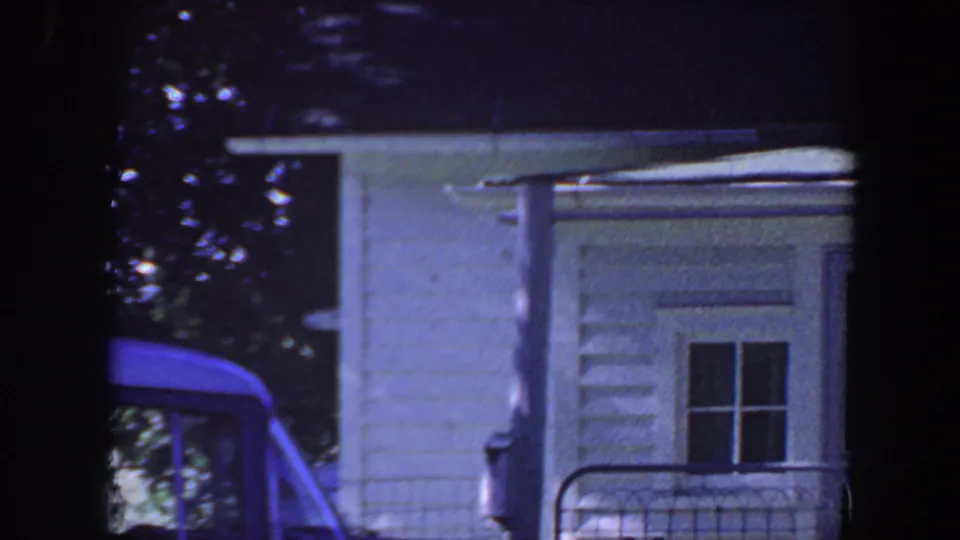 1959 Iowa home-movie frame showing a white house, porch railing, leafy trees, and part of a blue car.