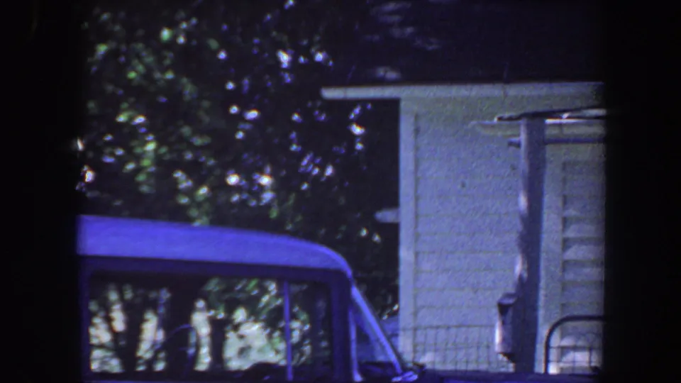 1959 Iowa 8mm frame with a blue car in the foreground, white house siding, porch railing, and shaded trees.