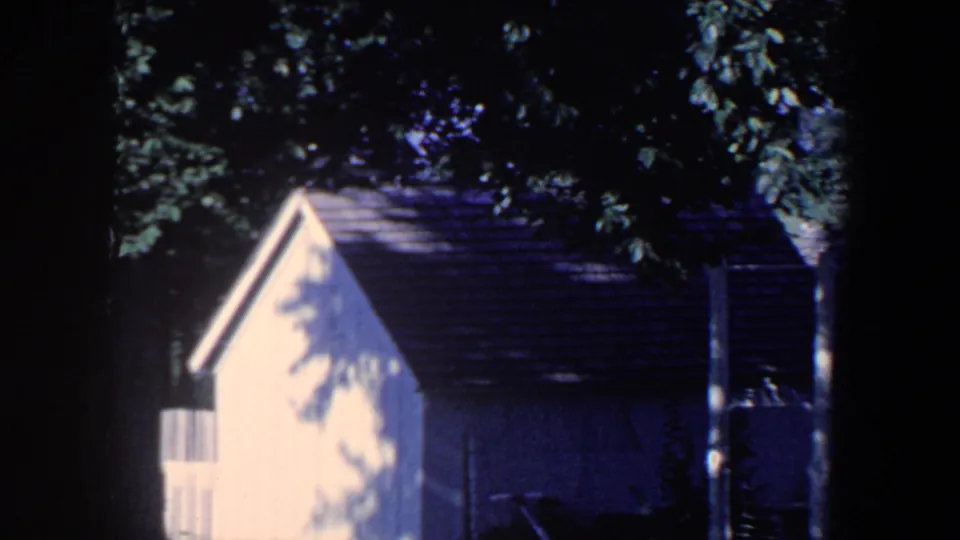 1959 Iowa home-movie frame showing a small yellow house, dark roof, tree shade, and backyard foliage.