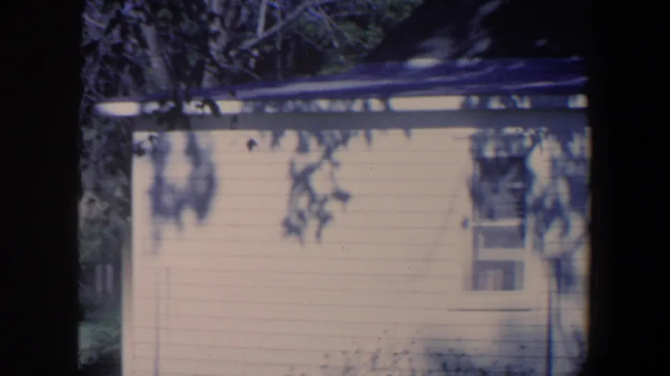 1959 Iowa home-movie frame centered on a pale outbuilding wall with a small window and tree shadows.