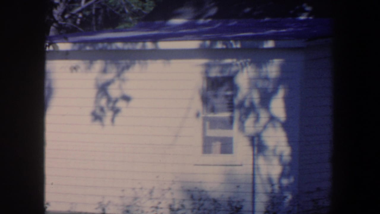 1959 Iowa 8mm frame showing dappled tree shadows on the side of a white house with a small window.