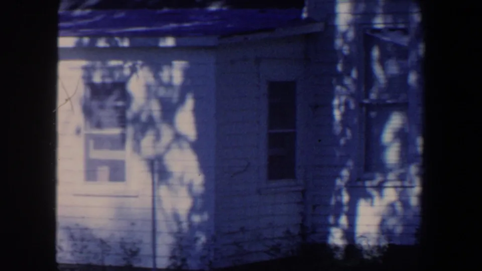 1959 Iowa home-movie frame of a white house exterior with windows and leaf shadows across the siding.