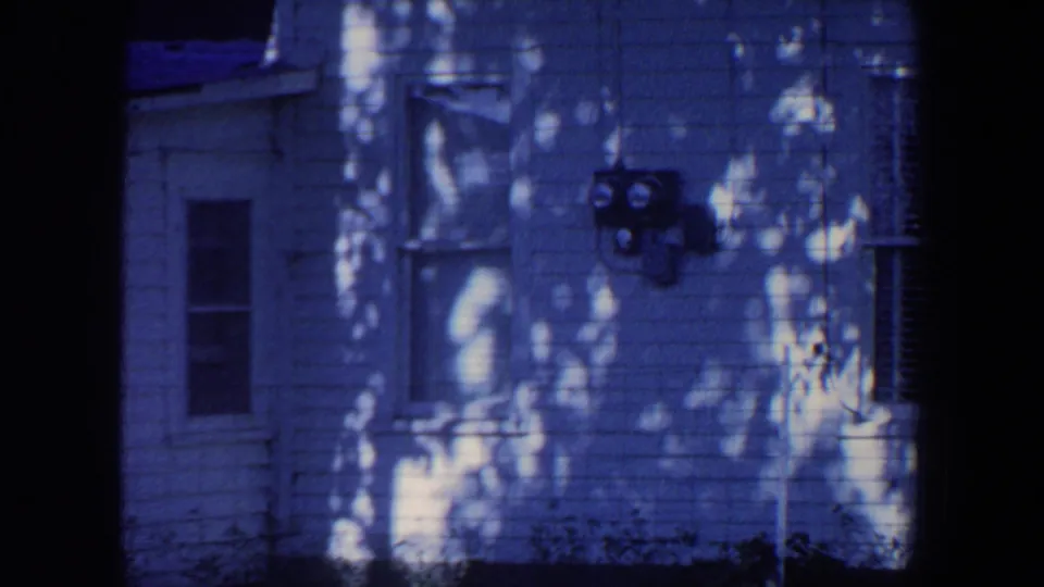 1959 Iowa 8mm frame showing a residential exterior with utility meters, a window, bushes, and dappled shade.