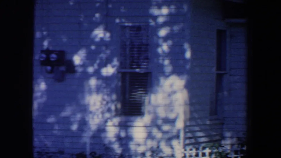 1959 Iowa frame of a white house wall with a shuttered window, exterior meters, and bright leaf shadows.