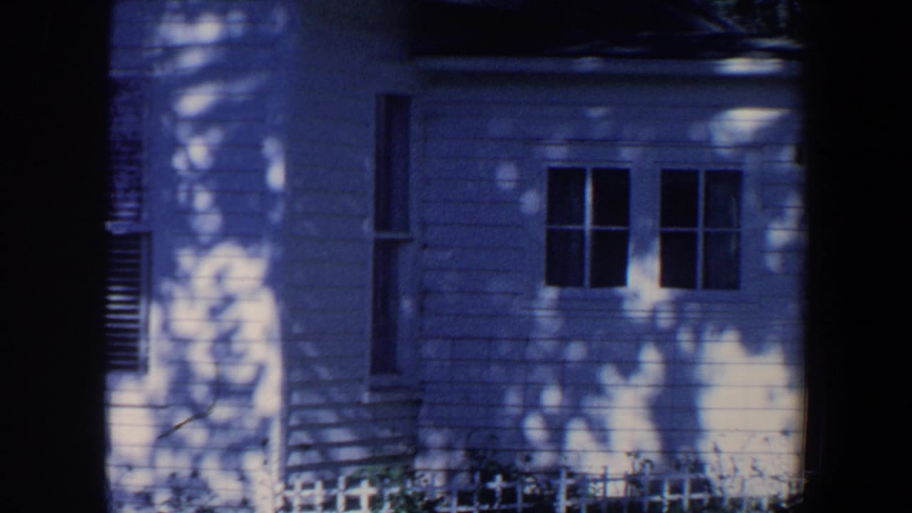 1959 Iowa home-movie frame showing a white clapboard house exterior, dark window openings, dappled tree shadows, and a low picket fence.