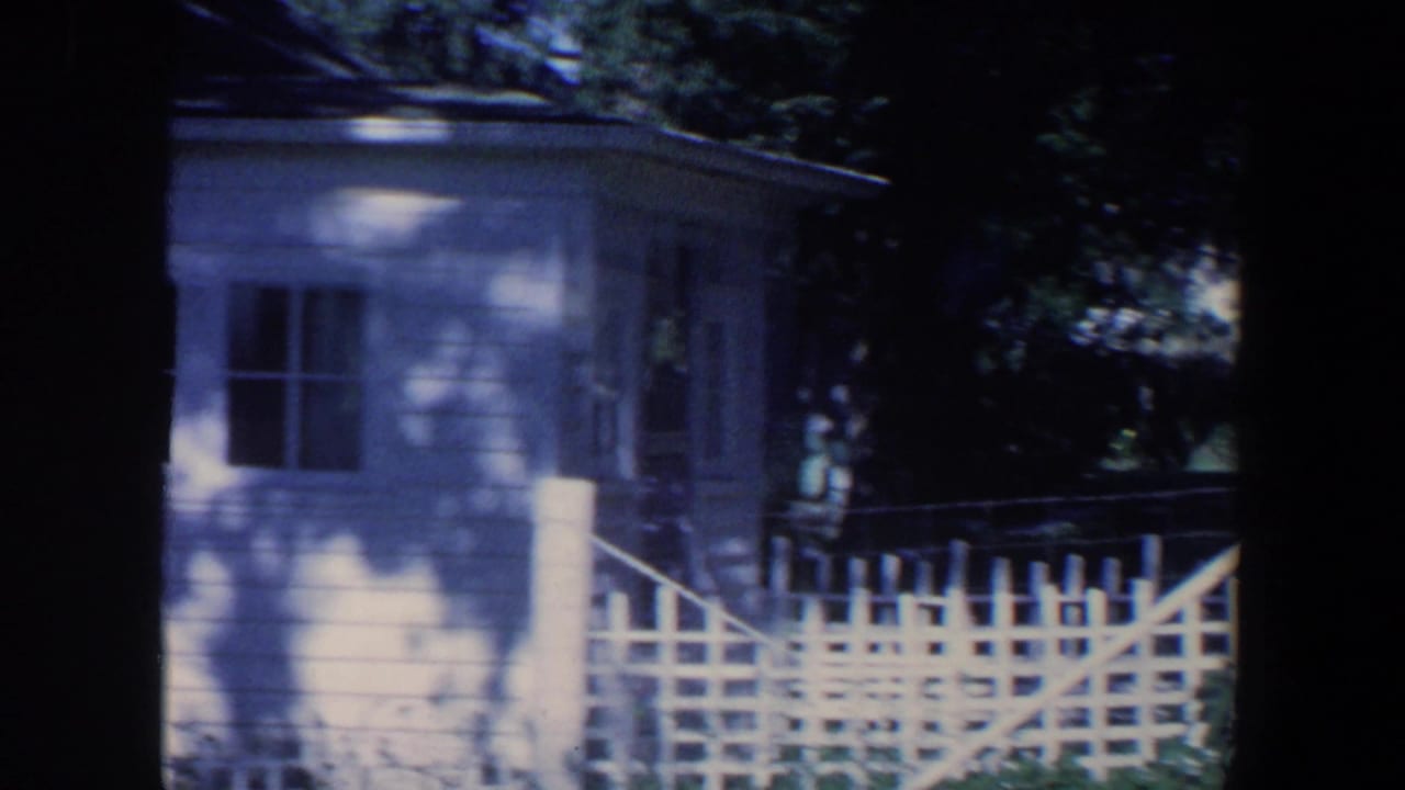 House corner with windows, picket fence, porch railing, and tree shade.