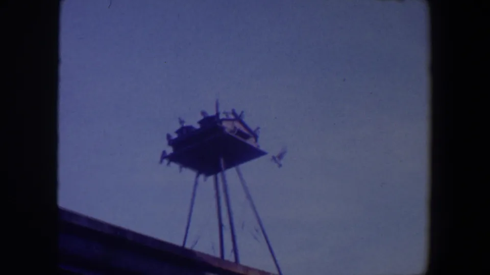 1959 Iowa 8mm frame showing birds gathered on a high wooden platform supported by thin metal legs against a blue sky.
