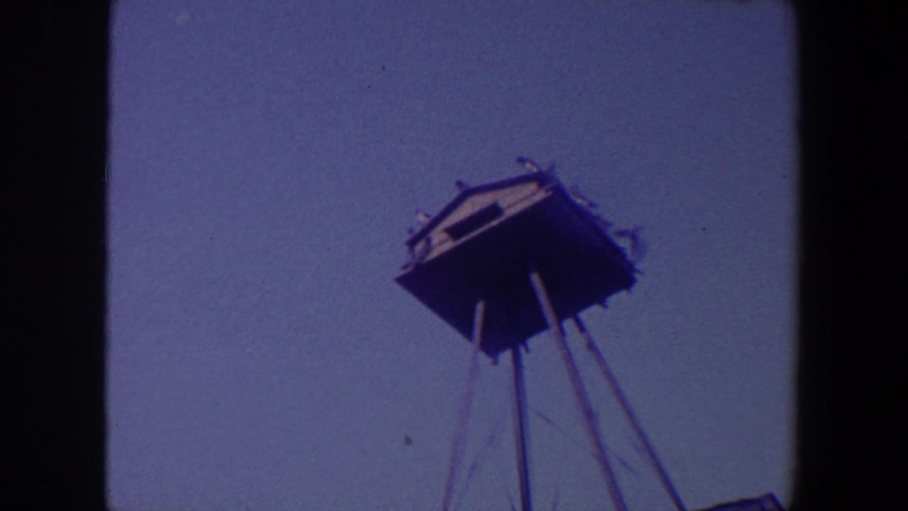 1959 Iowa 8mm frame showing an unidentified elevated structure on thin stilts, filmed from below against a muted sky.