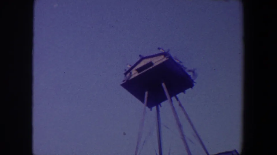 1959 Iowa 8mm frame showing an unidentified elevated structure on thin stilts, filmed from below against a muted sky.