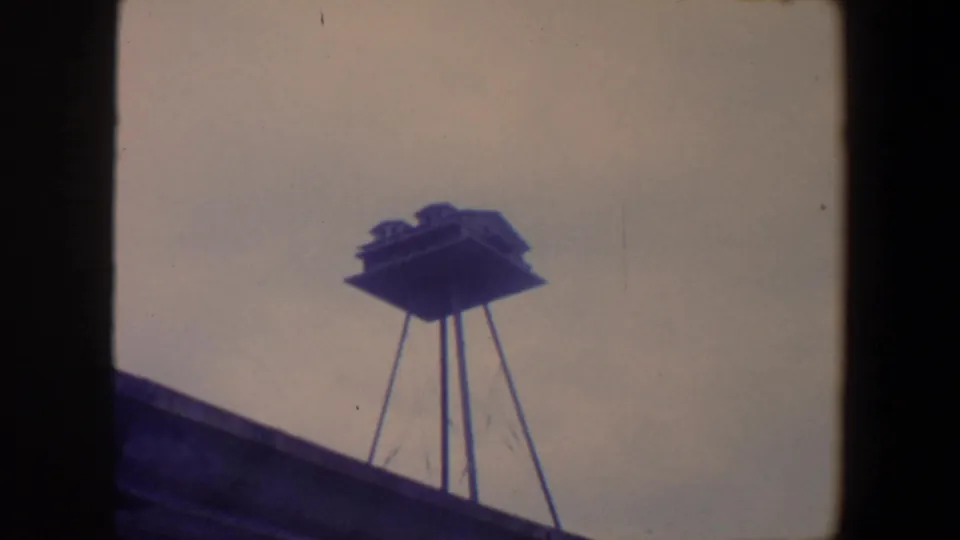 1959 Iowa 8mm frame with a raised structure on stilts seen above a dark roof edge or foreground building line.