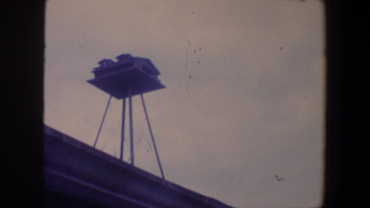 1959 Iowa 8mm frame showing a small raised birdhouse on slender supports above a roofline against a pale sky.