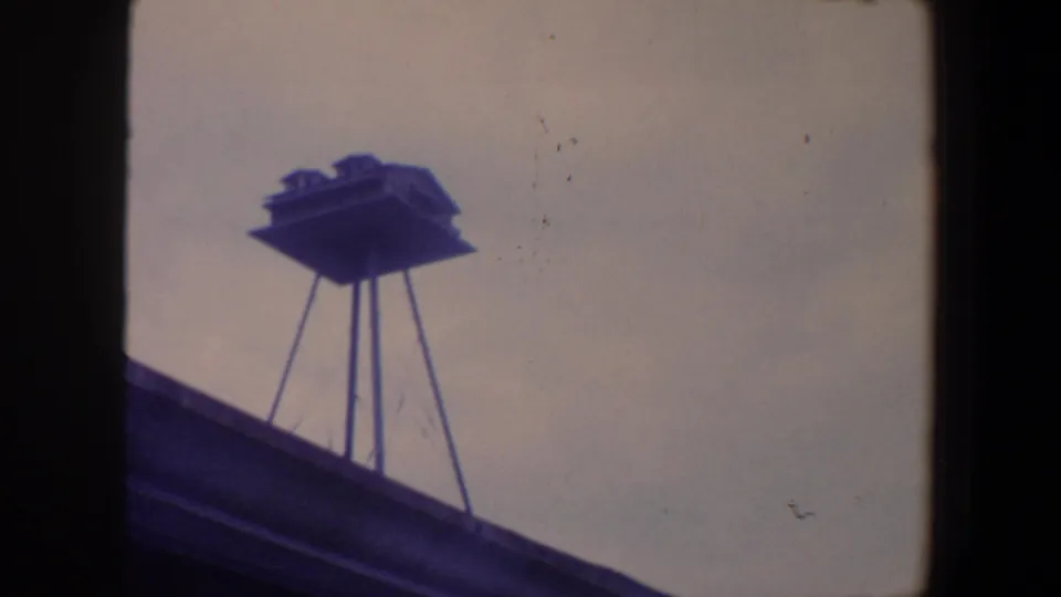 1959 Iowa 8mm frame showing a small raised birdhouse on slender supports above a roofline against a pale sky.