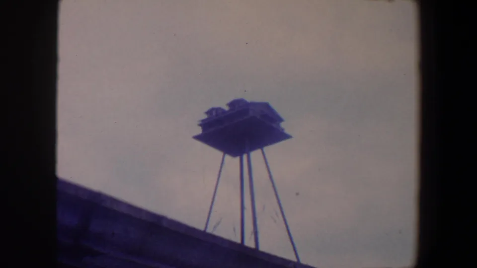 1959 Iowa home-movie frame with a raised rooftop birdhouse, thin supports, and cool-toned 8mm film texture.