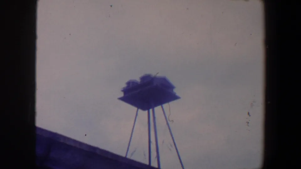 1959 Iowa 8mm frame of a small rooftop birdhouse on narrow supports, seen through scratches and cool color shift.