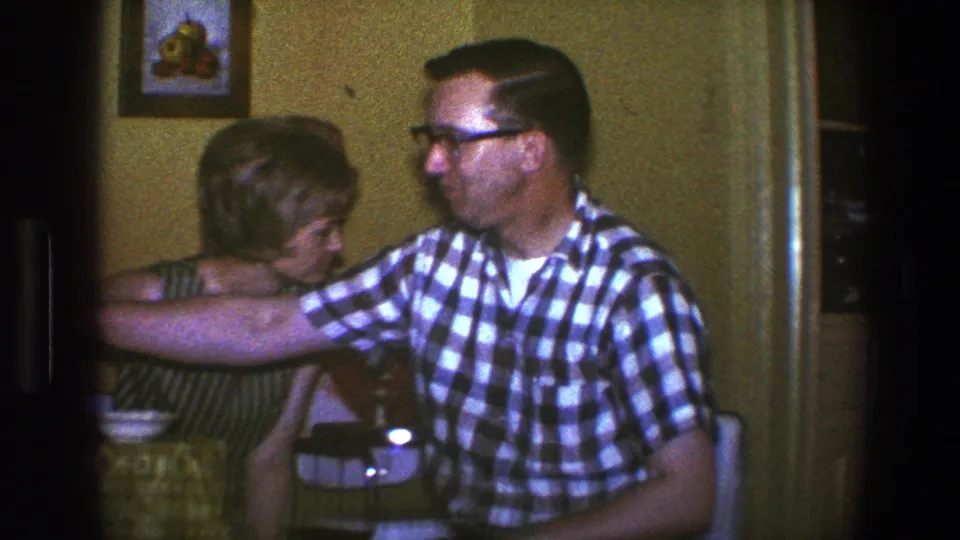1973 Saint Paul interior with a man in glasses reaching toward or past a seated woman near a table.