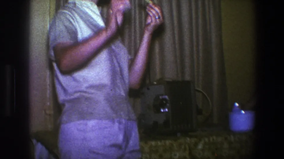 1973 Saint Paul interior showing a person standing near curtains, a table, and a possible film projector.