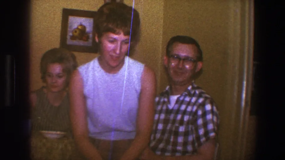1973 Saint Paul home-movie frame showing a blond woman seated indoors with a man in glasses and another adult partly visible behind her.