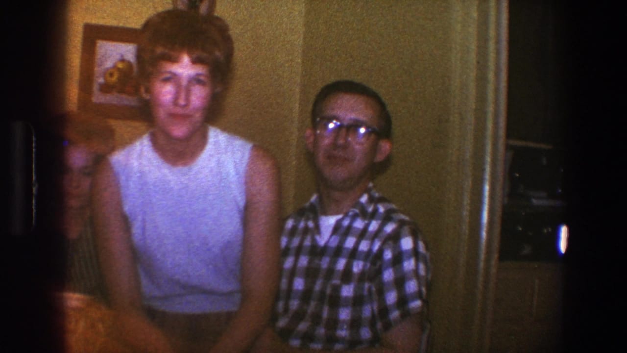 Blond woman faces the camera while a man in glasses sits beside her indoors.
