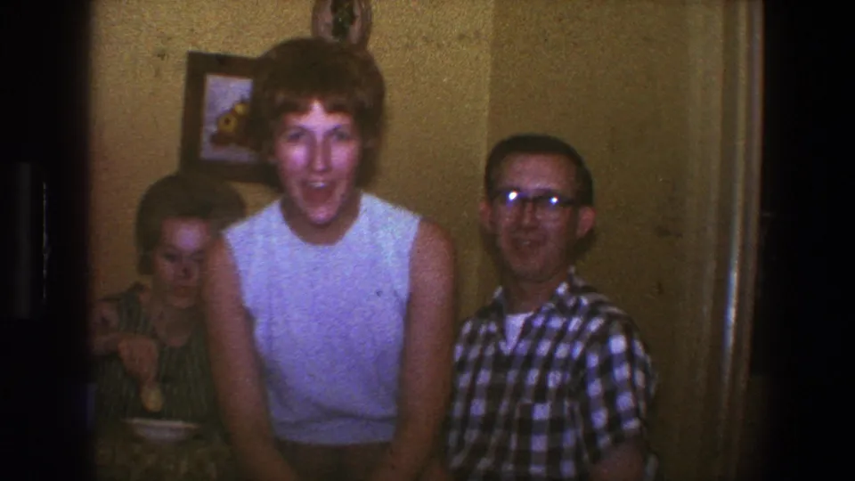 1973 Saint Paul frame showing a blond woman appearing to talk toward the camera, with a man in glasses and another adult nearby.