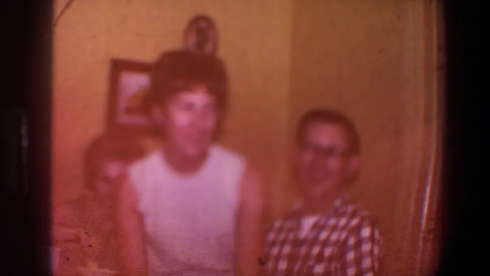 Soft 1973 Saint Paul home-movie frame with reddish exposure, showing seated adults indoors and the central woman blurred while reacting.