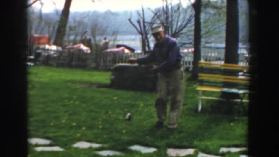 The man remains on the grass beside a painted bench, with a white fence and water visible behind him in the 1961 Novi footage.