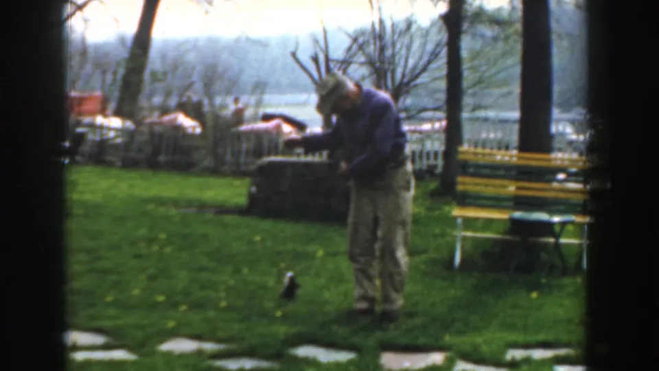 In Novi, Michigan in 1961, a man in a blue shirt bends over a fishing line on grass near a yellow-green bench, fence, trees, and lake.
