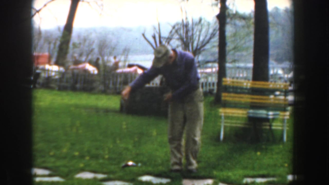 Man bends with arms lowered as a small fish lies on the grass near a bench and fence.