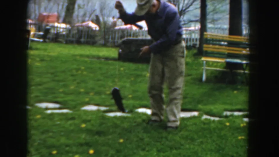 Near the end of the 1961 Novi clip, the man stands over the small fish on the grass with a yellow-green bench and lake visible behind him.