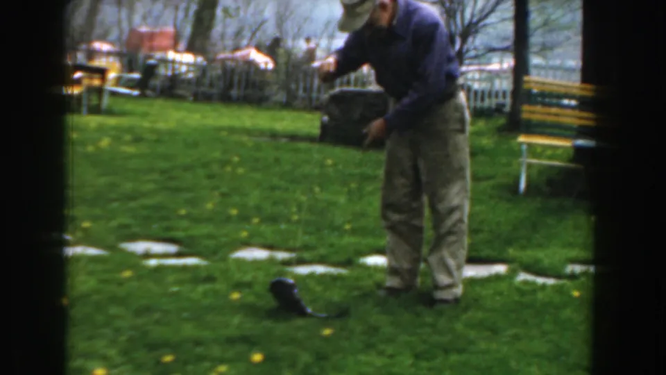 In 1961 Novi, Michigan, an older man stands on a green lawn holding a string attached to a catfish near stepping stones.