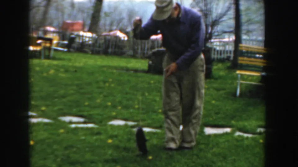 The man bends over the catfish on a Novi lawn, with a picket fence, bare trees, and a yellow-green bench behind him.