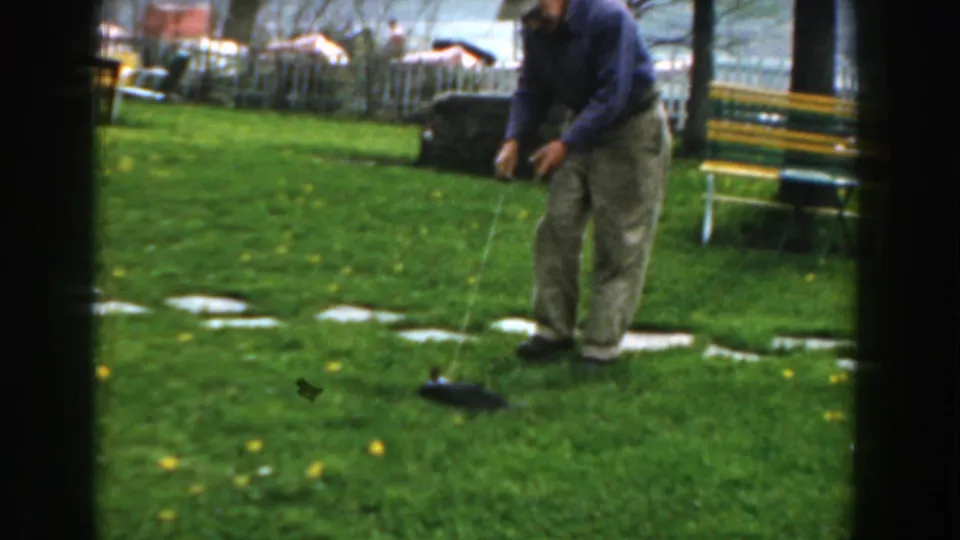 An older man in casual clothes lowers a string toward a catfish on a green backyard lawn in the Novi home-movie scene.