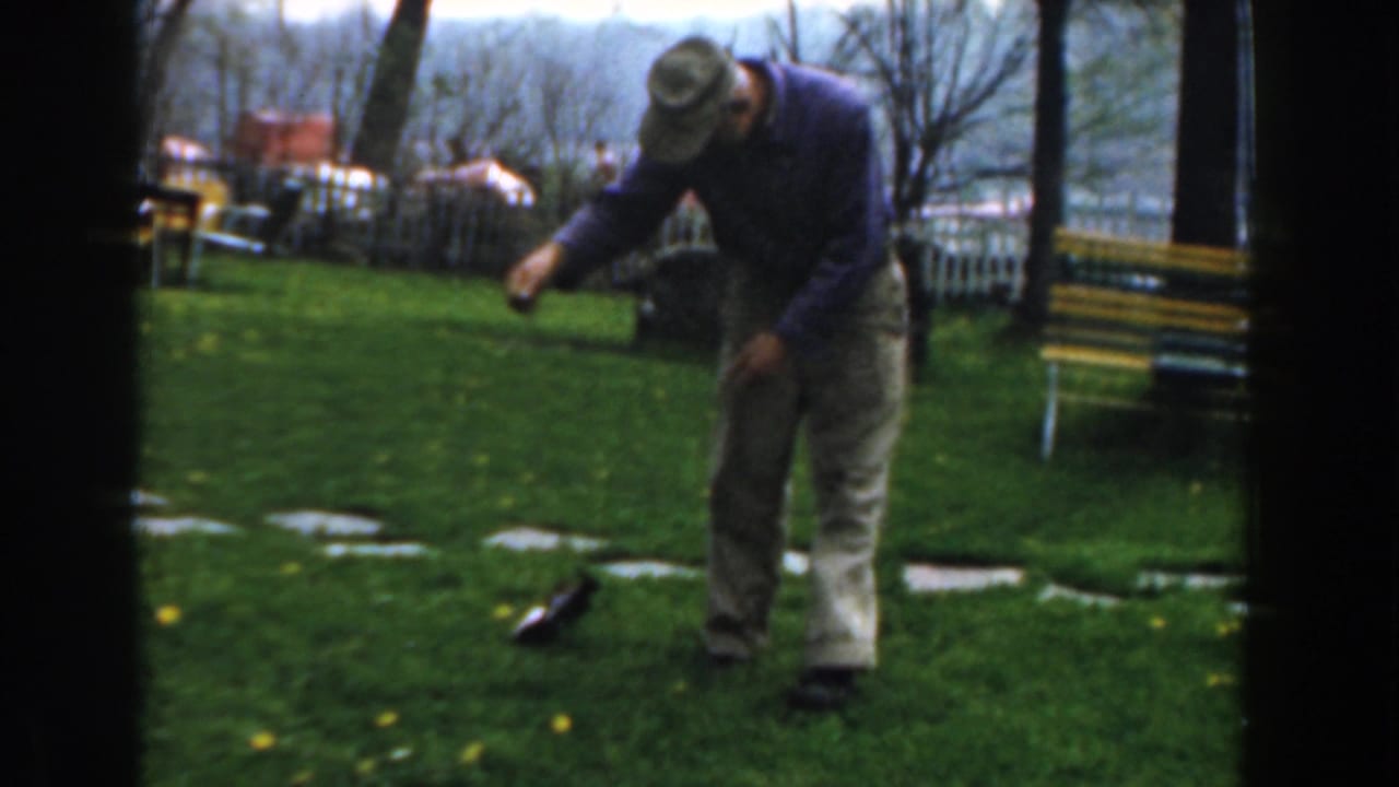 Man leans forward as a small fish lies or flips on grass near a bench and stone path.