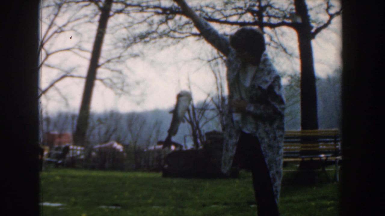 Woman in a patterned coat raises a pole with a small shark or fish in a park.